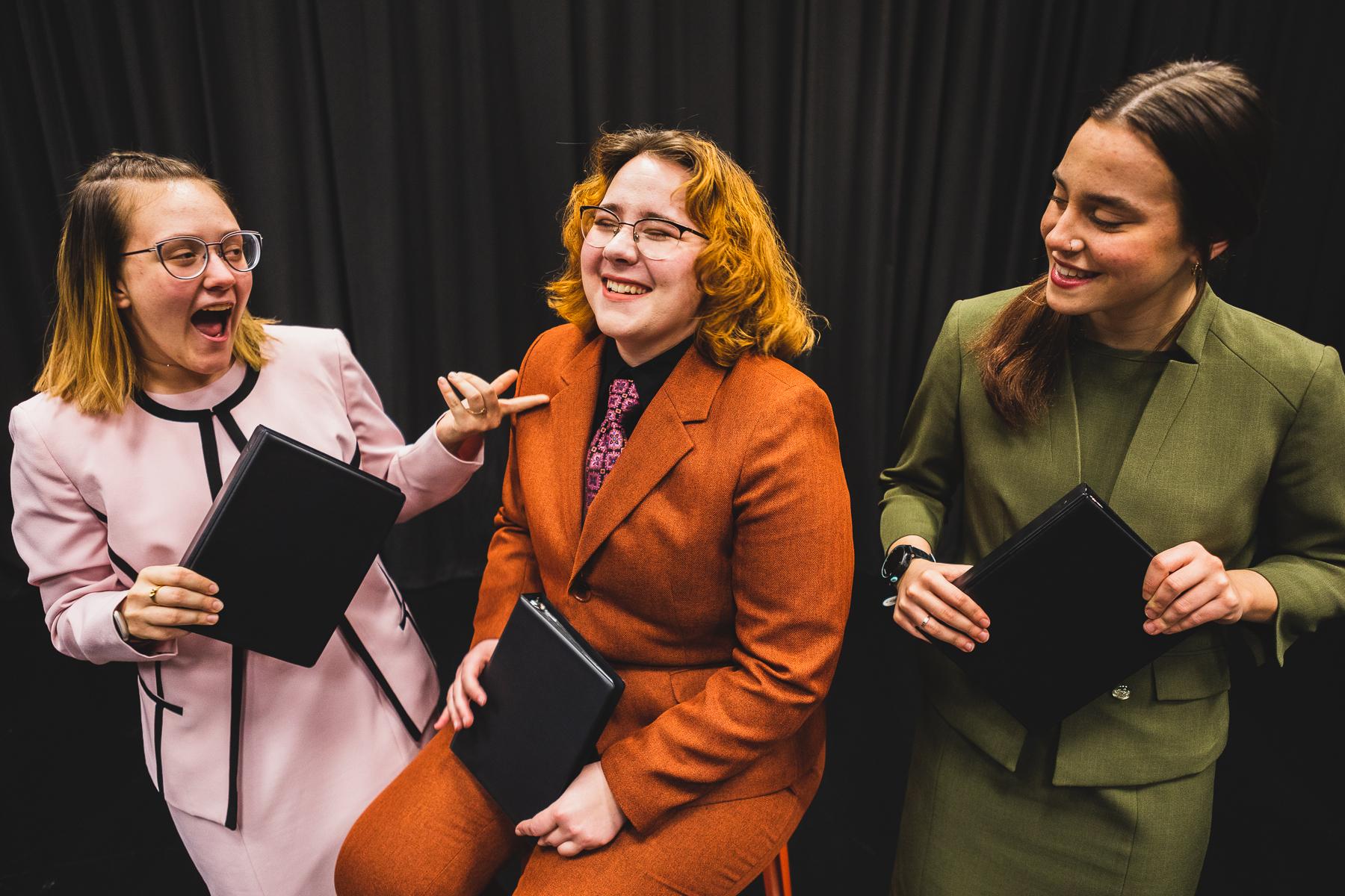 学生Maddy Ramey, wearing a green suit; Olivia Vore, wearing an orange suit with pink tie; and Ali Moulton, 穿着粉红色的西装, 拿着黑色的小活页夹，在黑色的窗帘前笑, 用于竞争性演讲中的解释性事件.